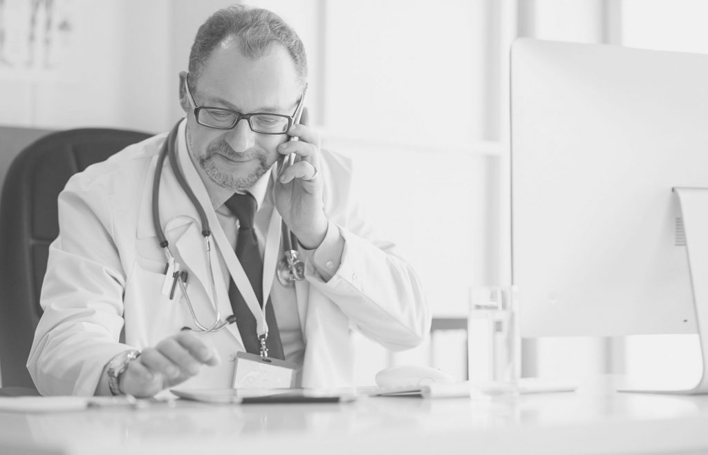 Zu sehen ist ein Arzt am Mobiltelefon, der gleichzeitig ein Tablet und einen iMac vor sich hat.